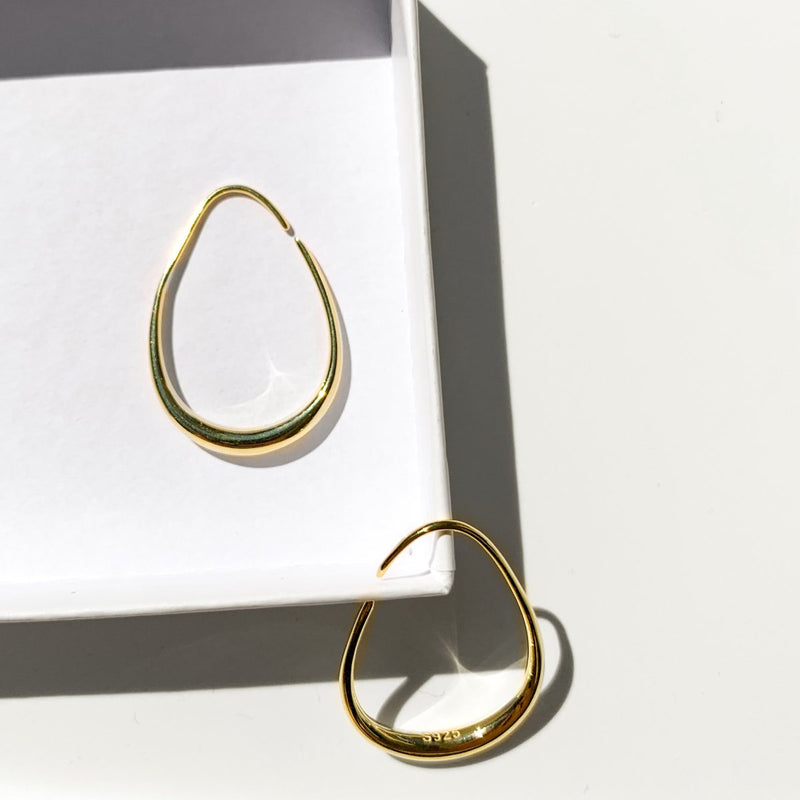 Golden Oval ear hoops lie in a product box, the hoops are open backed, thin at the top for easily wearing, and chunky at the bottom to really show off the style and design